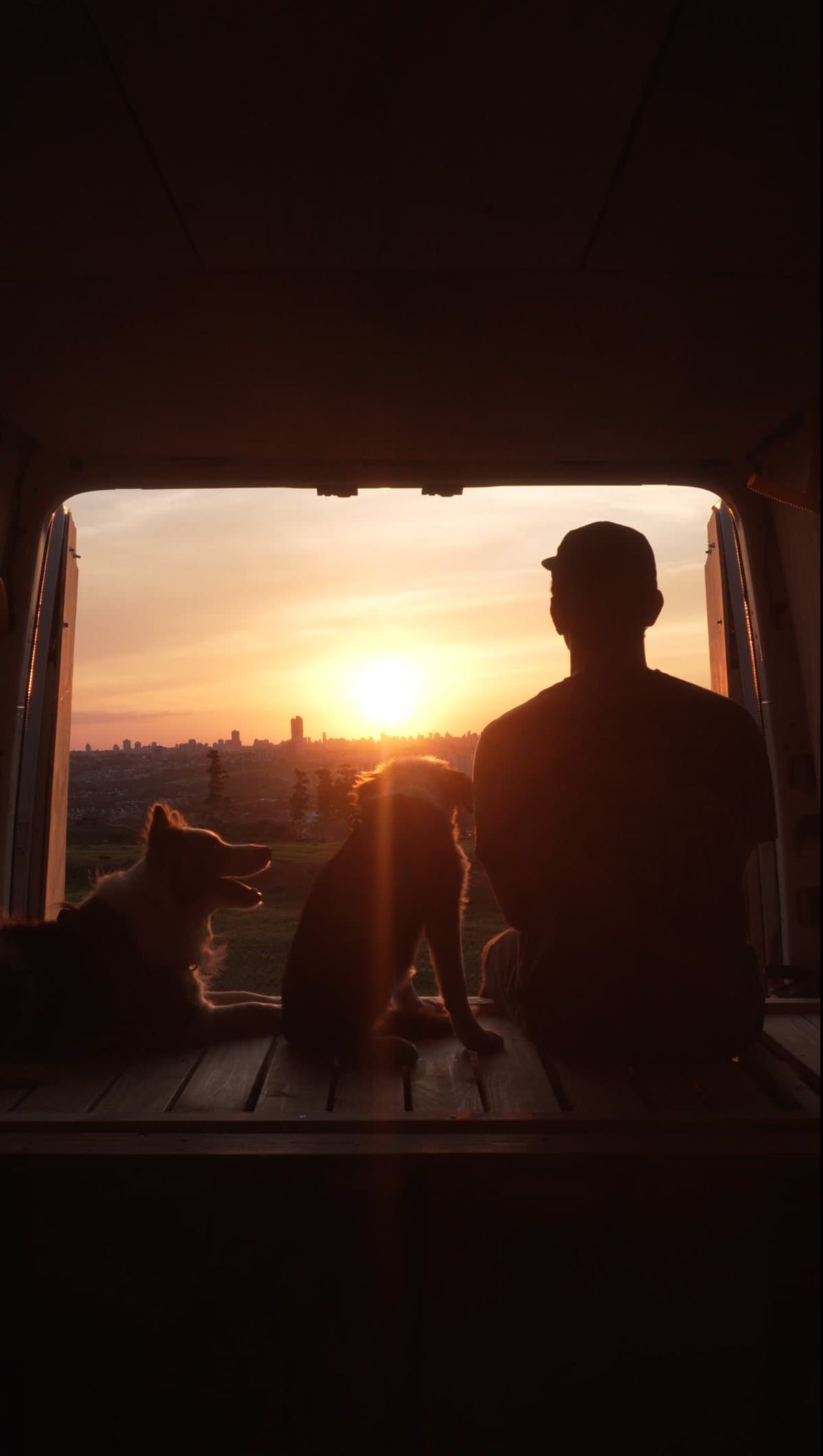 Daniel de costas na porta do motorhome, com as cachorras Bisteca e Panda, olhando o pôr do sol